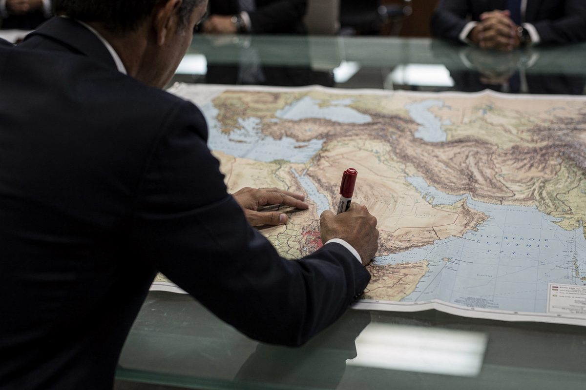 An over-the-shoulder view of a man's hands in a dark suit pressing a thick red marker pen heavily against a sprawling topographic map of the Arabian Peninsula.