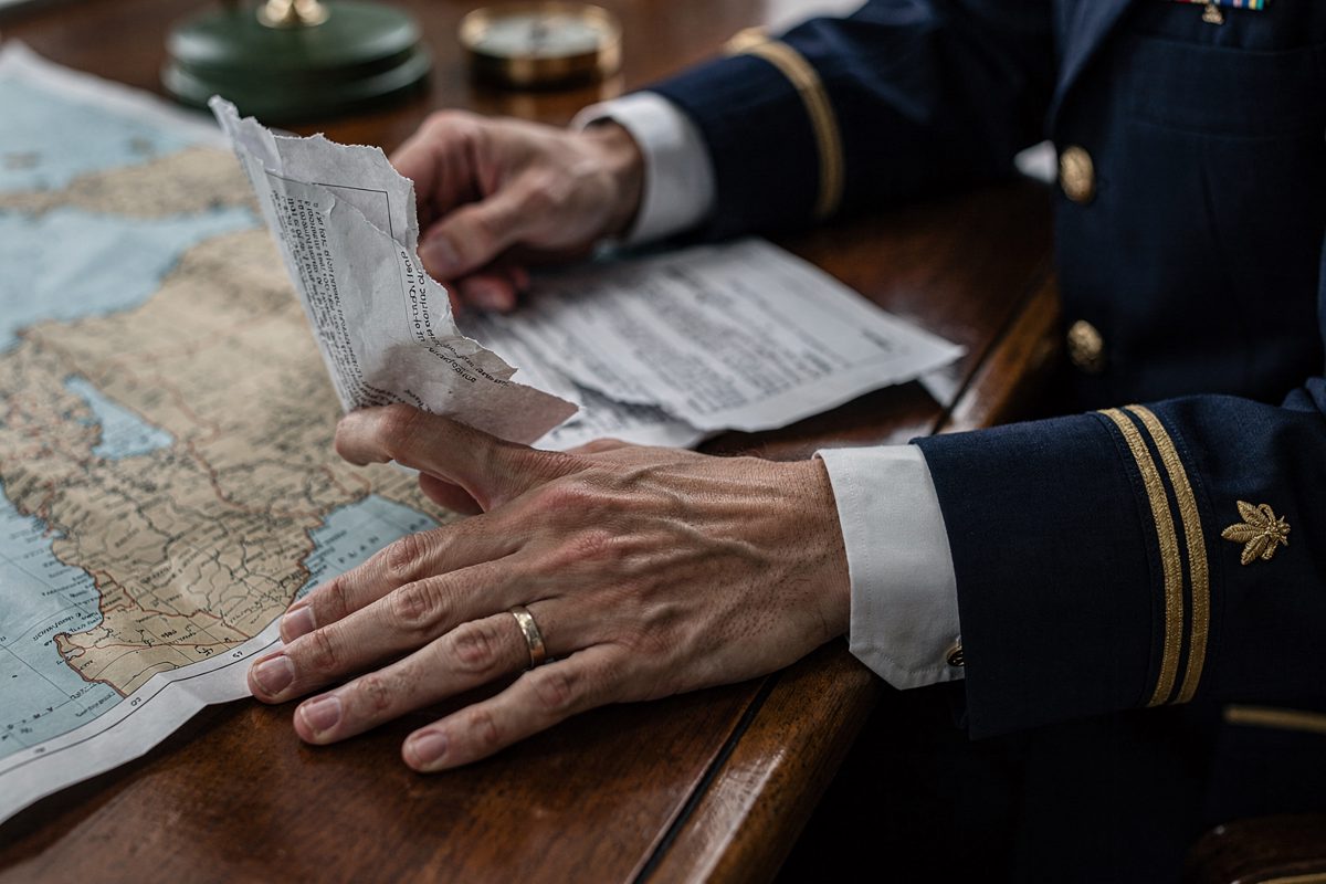 A close detail of hands in a crisp naval uniform resting on a polished wooden desk, with the fingers of one hand gripping a crumpled printed memorandum.