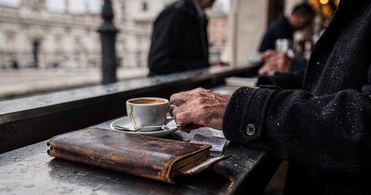 Close detail at a zinc café counter near the Vatican walls in soft early-morning light, a man's hand resting on a closed leather notebook beside a half-drunk.