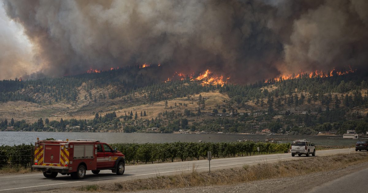 Wide documentary photograph from a roadside showing a distant forest wildfire northwest of a small British Columbia community, heavy smoke across the sky.