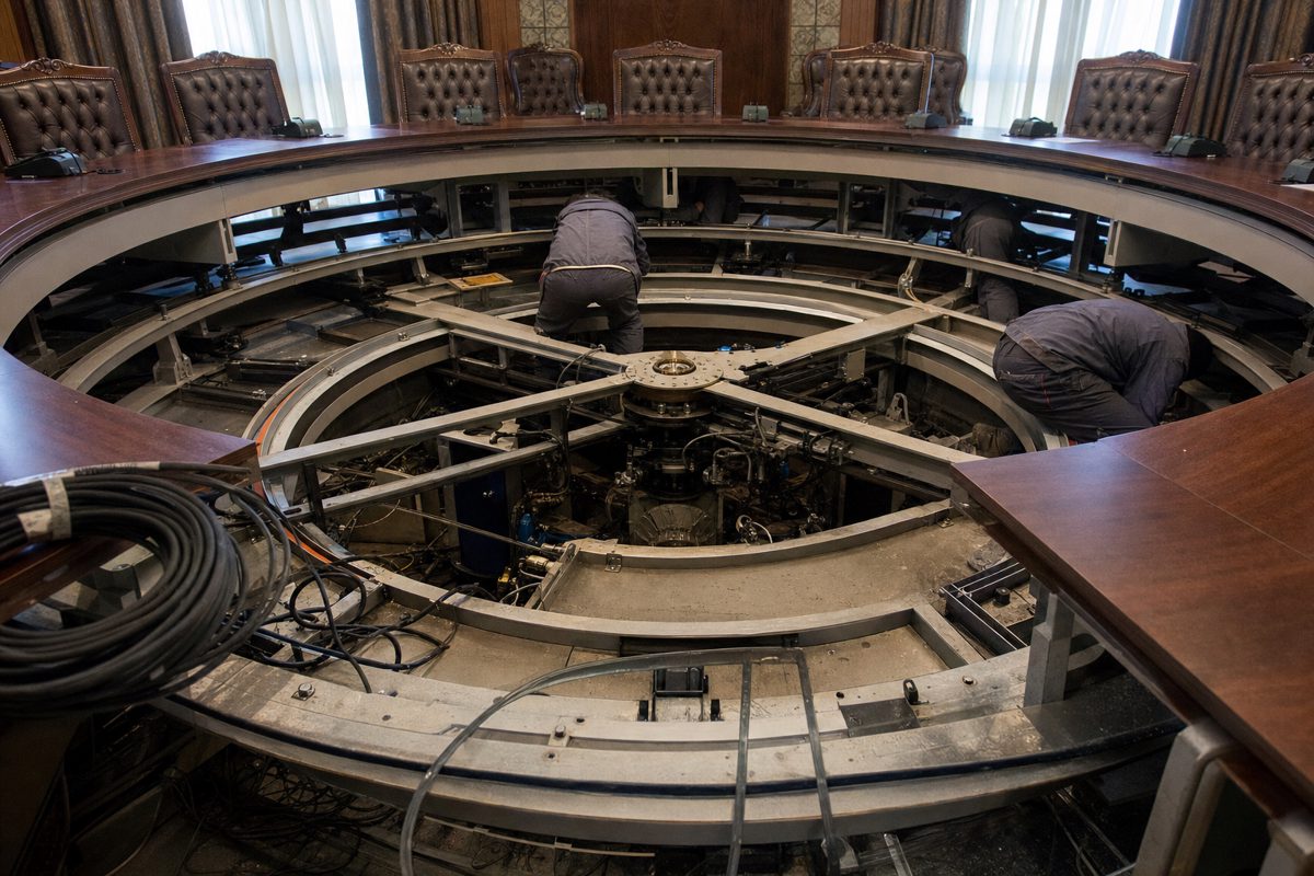 Documentary-style photograph of anonymous technicians inspecting the concealed mechanical base beneath a formal circular conference table, tools and cables.