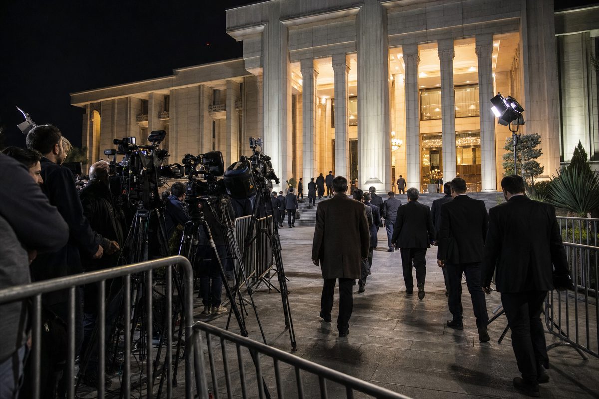 Night-time press photograph outside a monumental government building in Tehran, barriers, television cameras and anonymous officials entering beneath bright.