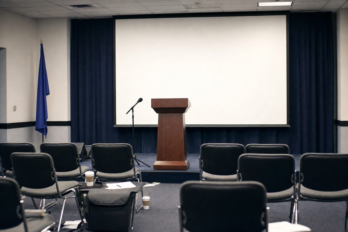 A generic government press-conference room: an empty podium bearing no insignia, a large blank projector screen behind it, rows of empty chairs and a scatter.