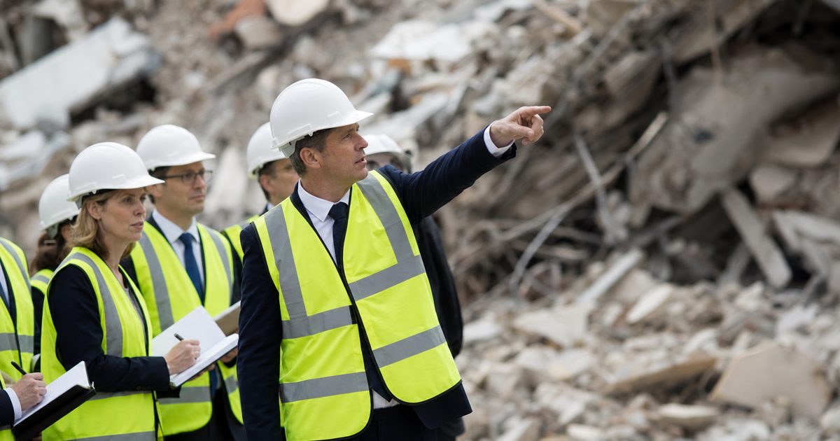 A formally dressed politician in a pristine, bright yellow high-visibility vest and a white hard hat stands awkwardly in front of an out-of-focus pile of.