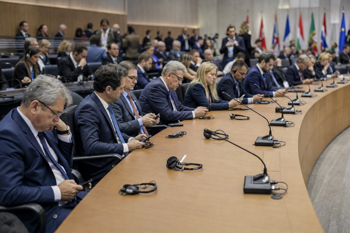 A wide shot of a sterile, modern international conference room. A row of diplomats in dark suits sit behind microphones at a long curved table, looking.