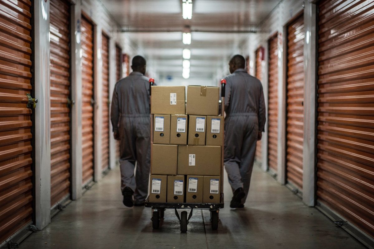 Wide shot down a long self-storage corridor under flat fluorescent light at first light, two workers in overalls seen from behind wheeling a trolley stacked.