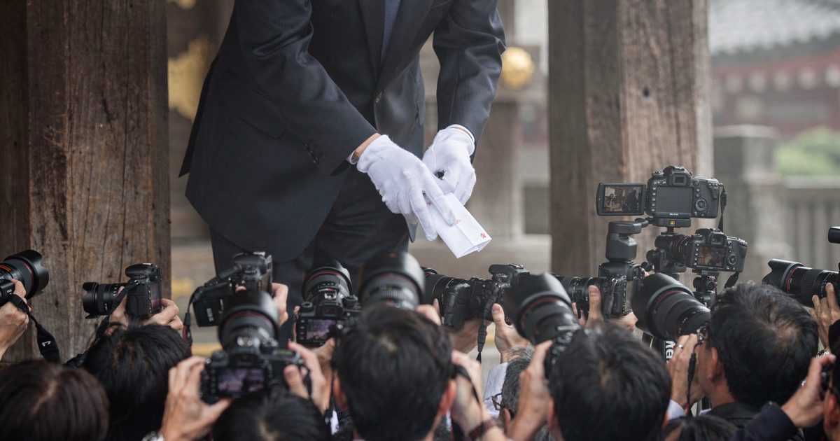 Close-up in hazy early-morning summer light at the wooden entrance gate of a Shinto shrine, the white-gloved hands of a suited official pausing mid-bow while a.