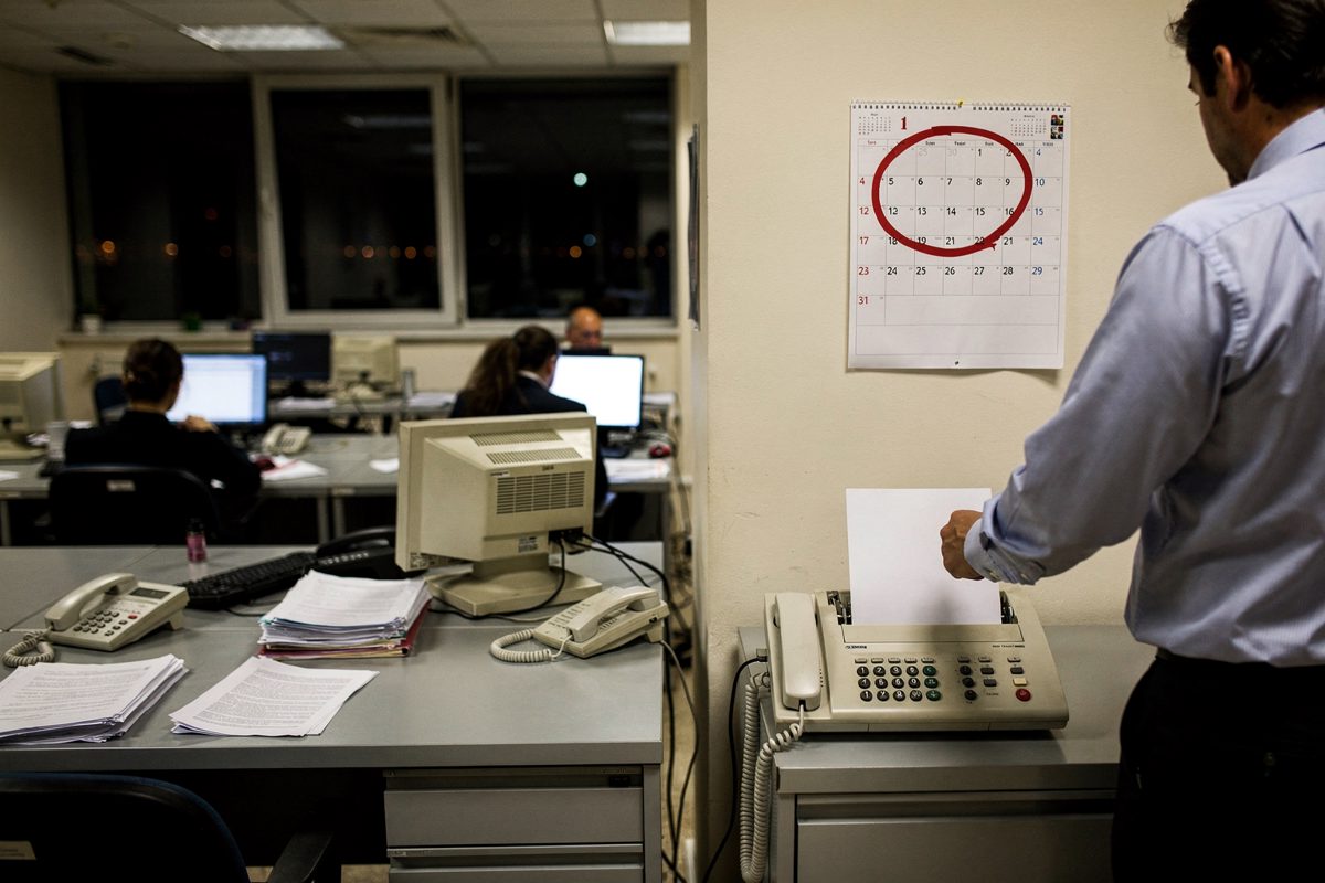 Wide interior shot of a fluorescent-lit foreign ministry office late at night, rows of desks seen from behind with one shirt-sleeved staffer part-cropped at.