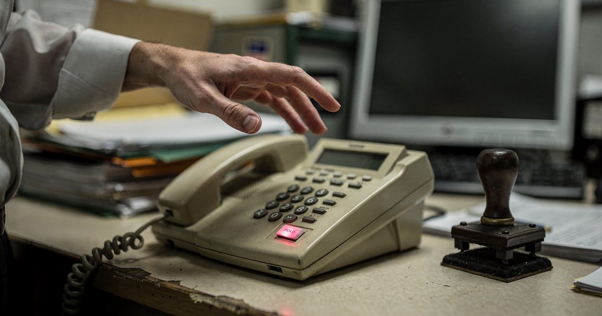 Close detail under flat office fluorescent light in a Treasury Department back office, a hand hovering over a beige desk telephone whose hold button blinks red.