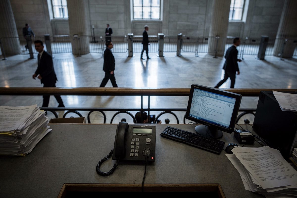 High angle from a mezzanine looking down into a marble federal lobby in Washington in cool early-morning light, a scatter of staff crossing the floor below.