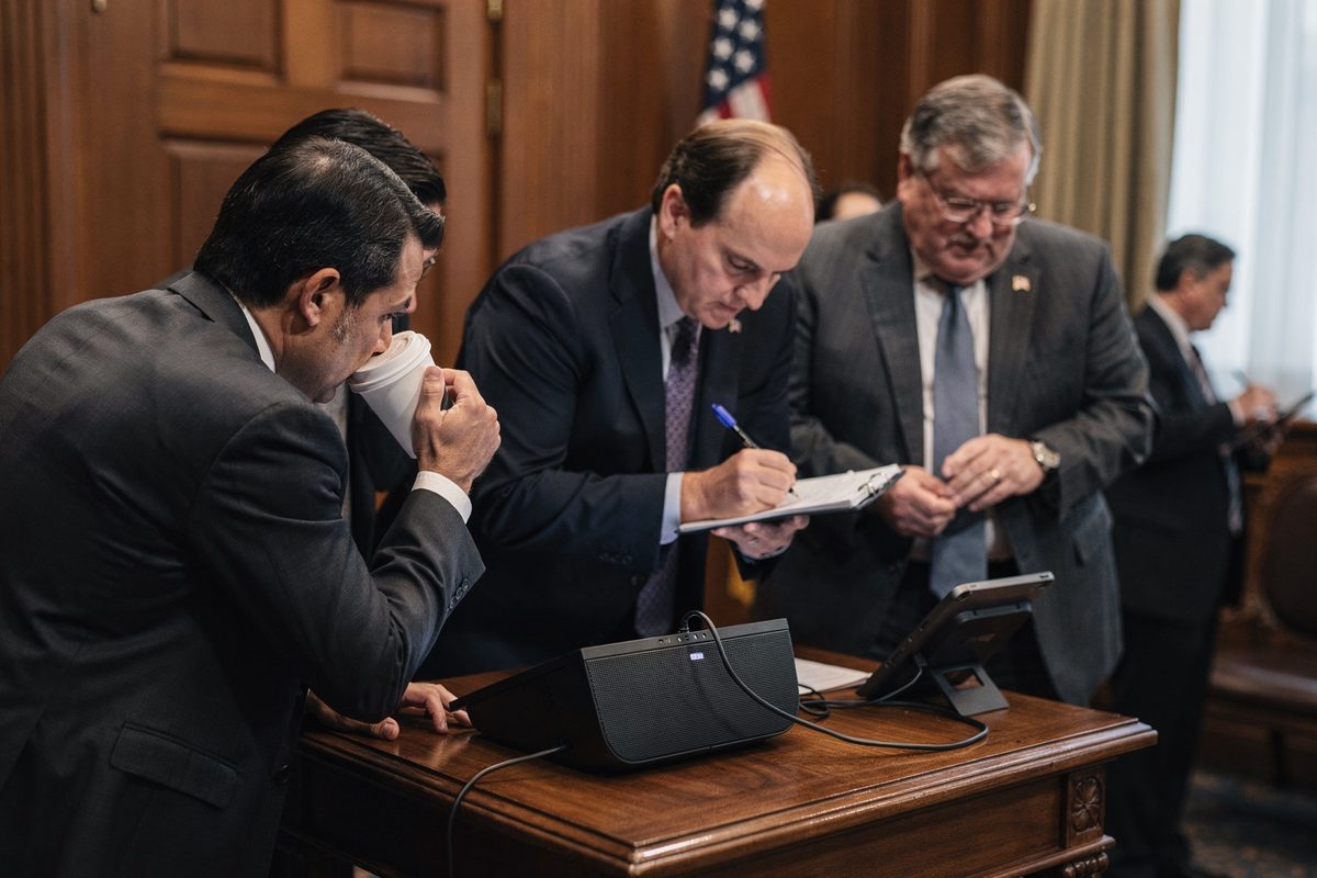 Medium shot from behind in a wood-panelled Senate committee anteroom at midday, half a dozen suited staffers leaning in around a conference speakerphone, one.